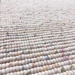 Puede incluir: Primer plano de una alfombra blanca texturizada con un patrón repetitivo de cuerdas gruesas y redondeadas. La alfombra tiene motas de color, incluyendo rosa, azul, amarillo y verde, que añaden interés visual. La alfombra parece estar hecha de una fibra natural suave.