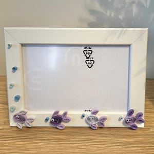 Peut inclure: Cadre photo rectangulaire blanc orné de poissons en papier roulé de couleurs violettes et bleues. Le cadre a un design épuré, avec de petits accents bleus et des ornements de poissons.