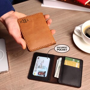May include: A tan leather wallet with visible stitching and the monogram TRH embossed on the front. The open wallet displays a clear ID window labeled document pocket, a vertical slot containing paper currency, and a black card slot.