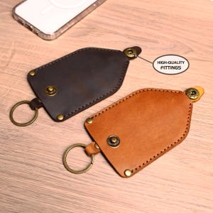 May include: Two leather keychains in brown and black, with brass hardware and stitching. The keychains have a snap closure and a metal ring for keys. The text "HIGH-QUALITY FITTINGS" is visible.