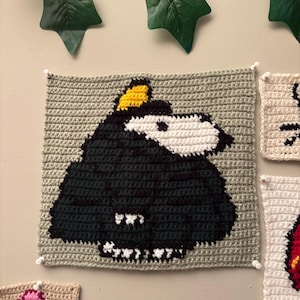 May include: A crocheted square featuring a black and white cartoon toucan with a yellow beak and head feathers. The toucan is set against a gray background. The square is part of a collection of crocheted art pieces.