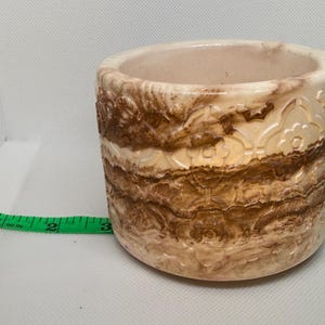 May include: A cylindrical stone planter with a cream and brown marbled finish featuring an embossed floral pattern. A green measuring tape is positioned in front, showing the base diameter is approximately 3.5 inches wide.