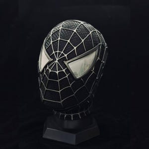 Op de afbeelding: Een zwart Spider-Man-masker met zilveren webdetails en reflecterende ooglenzen. Het masker wordt getoond op een zwarte standaard tegen een donkere achtergrond, wat het iconische superheldenontwerp laat zien.
