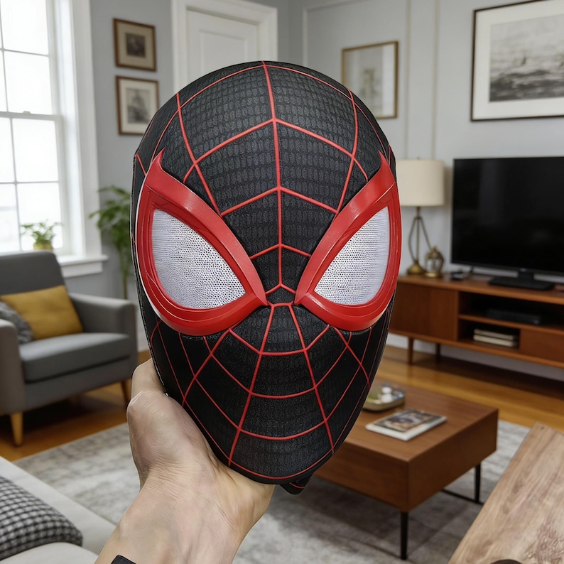 May include: A black and red Spider-Man mask with a web pattern. The mask has red eye frames and white mesh eye coverings. The mask is held up in front of a person in a living room setting.