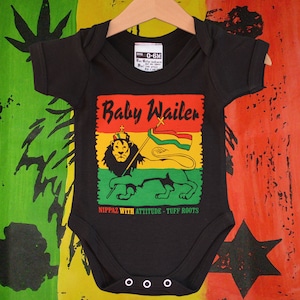 Puede incluir: Body de bebé negro con un gráfico colorido que presenta un león sosteniendo una bandera, las palabras "Baby Wailer" y el texto "Nippaz With Attitude - Tuff Roots". El body está colgado en una percha de madera.