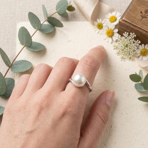 May include: A silver ring featuring a large, round, white pearl. The ring's band has a flowing, wave-like design. The ring is displayed on a finger, with a soft, neutral background and floral accents.
