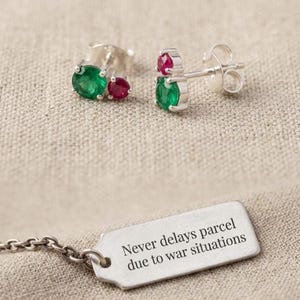 Puede incluir: Dos pendientes de botón con engaste de plata, cada uno con una esmeralda verde y un rubí rojo. También se ve una etiqueta plateada con el texto "Never delays parcel due to war situations".