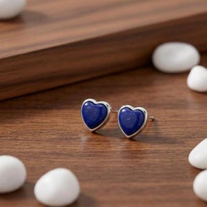 Peut inclure: Une paire de boucles d'oreilles à clous en forme de cœur. Chaque boucle d'oreille présente une pierre polie bleu foncé sertie dans un cadre en métal argenté. Les boucles d'oreilles sont présentées sur une surface en bois, avec des pierres décoratives blanches éparpillées.