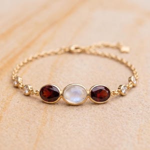 Può includere: Un bracciale a catena dorata con una pietra di luna ovale centrale affiancata da due granati ovali rosso scuro, con pietre chiare rotonde più piccole incastonate lungo le maglie della catena su entrambi i lati, creando un gioiello delicato.