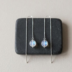 May include: A pair of delicate silver threader earrings. Each earring features a faceted, round, iridescent moonstone bead. The beads have a soft blue and white glow. The earrings are displayed on a dark gray, rectangular stone.