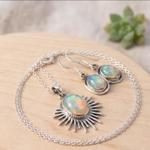 May include: A silver jewellery set consisting of a necklace and a pair of earrings. The necklace features an oval opal pendant with a sunburst design. The matching earrings have oval opal stones set in silver with simple hook-style backings.
