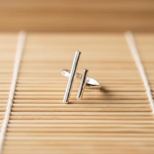 Op de afbeelding: Een zilveren ring met een minimalistisch design. De ring heeft twee verticale staven van verschillende lengtes, wat een moderne en geometrische esthetiek creëert. De ring staat tegen een lichtbruine, getextureerde achtergrond.