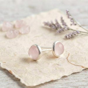 Könnte beinhalten: Ein Paar Ohrstecker mit glatten, runden, blassrosa Rosenquarz-Cabochons in polierten silberfarbenen Metallfassungen. Die Ohrstecker haben gerade Stifte und werden zusammen mit mehreren losen, getrommelten Rosenquarzsteinen präsentiert.