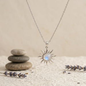 May include: A silver-toned necklace featuring a sun-shaped pendant with a central round iridescent moonstone. The pendant is suspended from a delicate silver-toned chain, showcasing radiating metal rays extending from the circular stone centre.