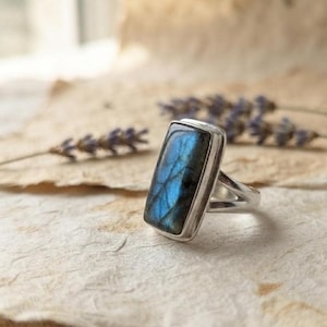 Puede incluir: Un anillo de plata con una piedra preciosa de labradorita rectangular de superficie pulida. La piedra muestra una iridiscencia azul vibrante con vetas oscuras naturales. La banda de metal es lisa y sencilla, sujetando la piedra en un engaste.