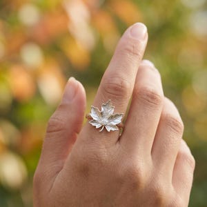 Può includere: Un anello d'argento con un design a forma di foglia d'acero dettagliato. L'anello presenta una piccola gemma trasparente al centro della foglia. L'anello è indossato su un dito, con uno sfondo sfocato di verde e arancione.