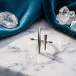 May include: A polished silver ring featuring two vertical bars of differing heights attached to a circular band. The metal surface is smooth and reflective, catching the light to create a bright, metallic sheen across the structure of the ring.
