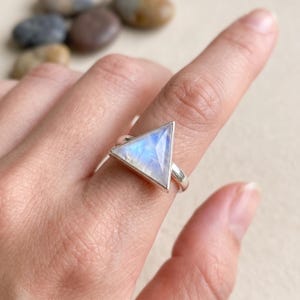 Pode incluir: Anel de prata com uma pedra da lua triangular iridescente, com tons de azul e branco. A pedra da lua é engastada numa banda de prata. O anel é mostrado num dedo.