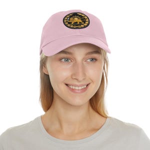 Op de afbeelding: Lichtroze baseballpet met een zwart, goud en geel embleem met een leeuw en zon. De pet is gemaakt van een zachte stof en heeft een gebogen klep. Het embleem is gecentreerd op de voorkant van de pet.