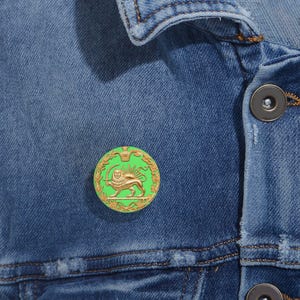 Puede incluir: Un pin redondo de color dorado con un fondo verde y un emblema de león. El pin está sujeto a una chaqueta vaquera azul. El león sostiene una espada y está rodeado por una corona dorada.