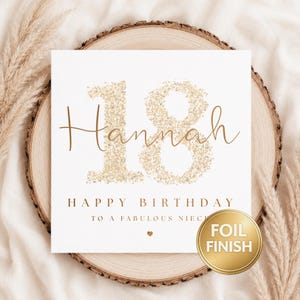 May include: A square white birthday card featuring the number 18 in a gold glitter texture. The name Hannah is written in gold script across the number. Below, the text reads HAPPY BIRTHDAY TO A FABULOUS NIECE with a small gold heart.