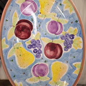 Könnte beinhalten: Eine ovale Keramikplatte mit einem handgemalten Obstmotiv auf hellblau gesprenkeltem Hintergrund. Das Design zeigt gelbe Birnen, dunkelrote Äpfel, violette Pflaumen und Traubenbündel mit grünen Blättern, die über die gesamte Fläche verteilt sind.