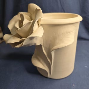 Könnte beinhalten: Eine beige Keramikvase mit einem geformten Rosen- und Blattdesign. Die Rose erstreckt sich von der Seite der Vase, und ein Blatt ist in die Seite geschnitzt. Die Vase hat eine abgerundete Oberseite und eine zylindrische Form.