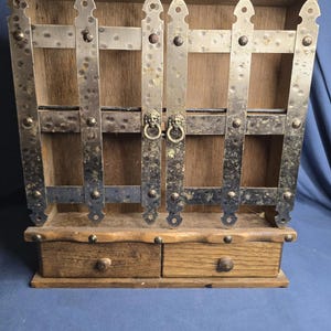 Könnte beinhalten: Ein hölzerner Wand-Schrank mit zwei kleinen Schubladen und einem dekorativen Metalltor-Design. Der Schrank hat eine natürliche Holzoberfläche mit Metallakzenten und eine geschwungene Oberseite. Das Metalltor hat Löwenkopfgriffe.