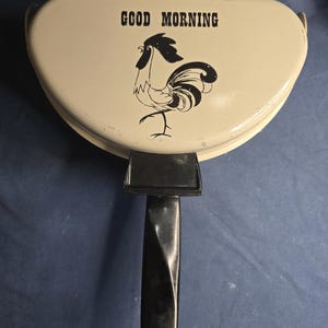 May include: Cream-colored, vintage pancake maker with a black handle. The lid features the words "GOOD MORNING" above a black rooster illustration. The handle is black and has a hole at the end for hanging.