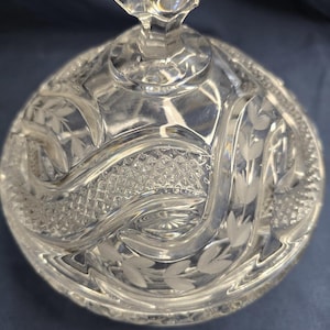 Peut inclure: Un plat à bonbons en verre transparent avec un couvercle surmonté d'un bouton à facettes. Le couvercle présente des motifs de feuilles gravées, des textures en losanges croisés et des bandes courbes lisses formant un motif répétitif.