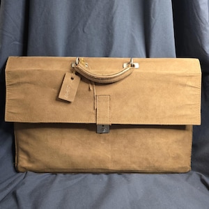 May include: A tan suede briefcase featuring a top handle, a rectangular flap closure with a central silver-toned metal lock, and a small rectangular luggage tag attached to the handle with a thin cord. The bag has a structured, flat design.
