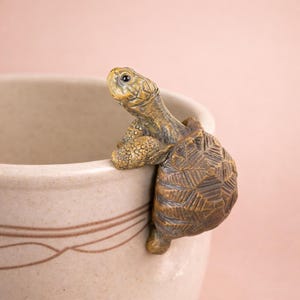 Puede incluir: Una figura de tortuga de cerámica marrón y beige, agarrada al borde de una taza de cerámica beige. La tortuga tiene un caparazón detallado y piel texturizada. La taza presenta un diseño sencillo con una línea marrón alrededor del borde.