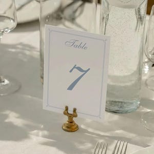 Puede incluir: Una tarjeta blanca con el número de mesa con la palabra «Table» y el número 7 en azul. La tarjeta se sujeta con un soporte de metal dorado. La mesa incluye cristalería y una botella de agua.