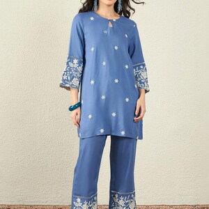 Könnte beinhalten: Ein zweiteiliges blaues Outfit, bestehend aus Tunika und Hose. Die Tunika hat einen Schlüsselloch-Ausschnitt und weiße Blumenstickereien an den Ärmeln und der Vorderseite. Die Hose hat passende Blumenstickereien an den Säumen. Das Outfit ist aus einem weichen, fließenden Stoff gefertigt.