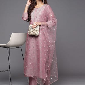 May include: A mauve-coloured, three-piece embroidered Indian suit. The set includes a long tunic, matching trousers, and a sheer dupatta with silver embellishments. The tunic has a round neckline and three-quarter sleeves. A small, embroidered clutch is held.