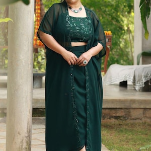 Peut inclure: Un ensemble trois pièces vert émeraude foncé composé d'un haut court sans manches avec une broderie ton sur ton complexe, une jupe taille haute assortie avec un drapé sur le devant et un long gilet transparent ouvert avec des bordures brodées.