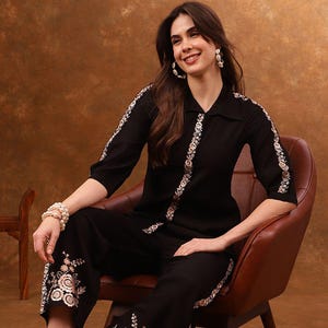May include: Black two-piece outfit with floral embroidery. The set comprises a collared shirt with three-quarter sleeves and wide-leg trousers. The floral embroidery is in cream and gold. The outfit is worn with black heels.