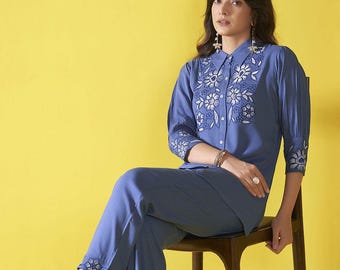 Puede incluir: Conjunto azul de dos piezas con bordados florales blancos. El conjunto consta de una camisa con botones y pantalones a juego. El conjunto se usa con tacones abiertos de color rosa claro. El fondo es amarillo liso.