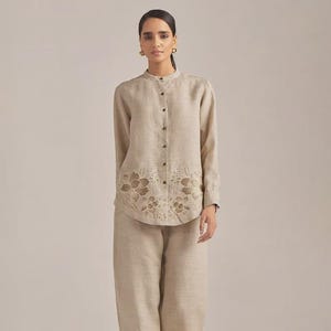 Puede incluir: Un conjunto de dos piezas de lino beige. La camisa abotonada de manga larga presenta un cuello mandarín y detalles florales recortados en el dobladillo. El conjunto incluye pantalones de pernera ancha a juego. El modelo lleva sandalias marrones.