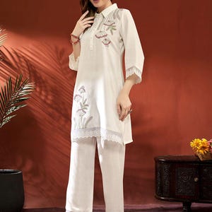 Puede incluir: Conjunto de dos piezas color crema. La parte superior es una t&uacute;nica con cuello, bordado floral y ribete de encaje, combinada con pantalones de pernera ancha a juego. El conjunto se lleva con sandalias de tac&oacute;n transparentes. El fondo es marr&oacute;n rojizo c&aacute;lido.