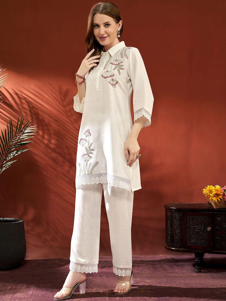 Puede incluir: Conjunto de dos piezas color crema. La parte superior es una t&uacute;nica con cuello, bordado floral y ribete de encaje, combinada con pantalones de pernera ancha a juego. El conjunto se lleva con sandalias de tac&oacute;n transparentes. El fondo es marr&oacute;n rojizo c&aacute;lido.