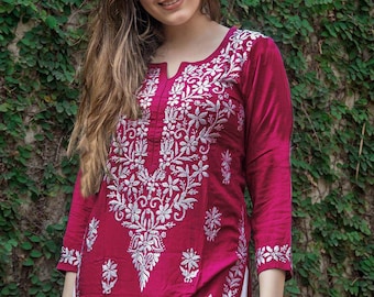 Op de afbeelding: Een magenta tuniek met gedetailleerde witte bloemenborduurwerk. De top met lange mouwen heeft een gekerfde halslijn en ingewikkelde patronen op de voorkant, mouwen en zoom. Het kledingstuk is gemaakt van een zachte, vloeiende stof.