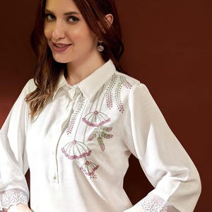 Puede incluir: Camisa blanca con bordado floral en rosa y verde. La camisa presenta detalles de encaje en las mangas y los pu&ntilde;os. El cuello es de estilo cl&aacute;sico y la camisa tiene un corte entallado.