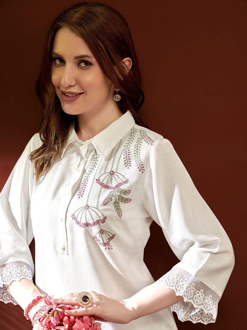 Puede incluir: Camisa blanca con bordado floral en rosa y verde. La camisa presenta detalles de encaje en las mangas y los pu&ntilde;os. El cuello es de estilo cl&aacute;sico y la camisa tiene un corte entallado.