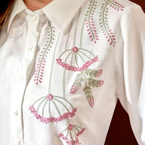 Puede incluir: Camisa blanca con bordado floral. El bordado presenta dise&ntilde;os florales rosas y verdes en la parte delantera y en el &aacute;rea del hombro. La camisa tiene cuello cl&aacute;sico y pu&ntilde;os abotonados.