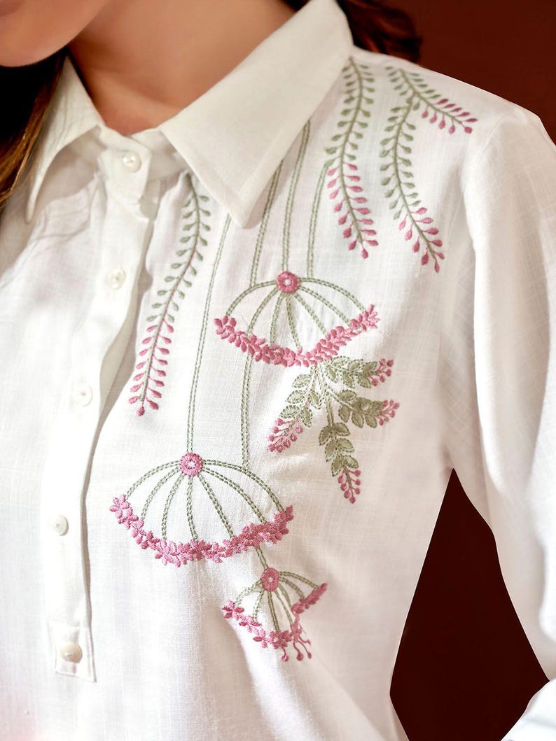 Puede incluir: Camisa blanca con bordado floral. El bordado presenta dise&ntilde;os florales rosas y verdes en la parte delantera y en el &aacute;rea del hombro. La camisa tiene cuello cl&aacute;sico y pu&ntilde;os abotonados.