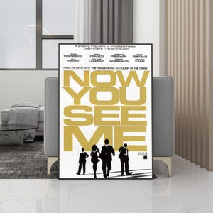Puede incluir: Un póster de película enmarcado de "Ahora me ves" con un marco negro. El póster presenta letras doradas y siluetas de personas. El texto incluye el título de la película, los nombres del elenco y el año 2013.