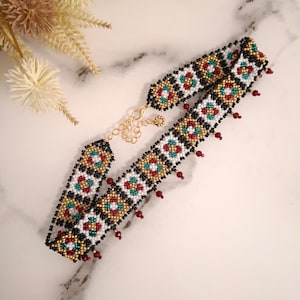 K&ouml;nnte beinhalten: Eine Halskette mit Perlen im Choker-Stil mit einem geometrischen Muster in Schwarz, Wei&szlig;, Gold, Rot und T&uuml;rkis. Die Halskette hat einen goldfarbenen Kettenverschluss und kleine rote Perlen, die unten h&auml;ngen.