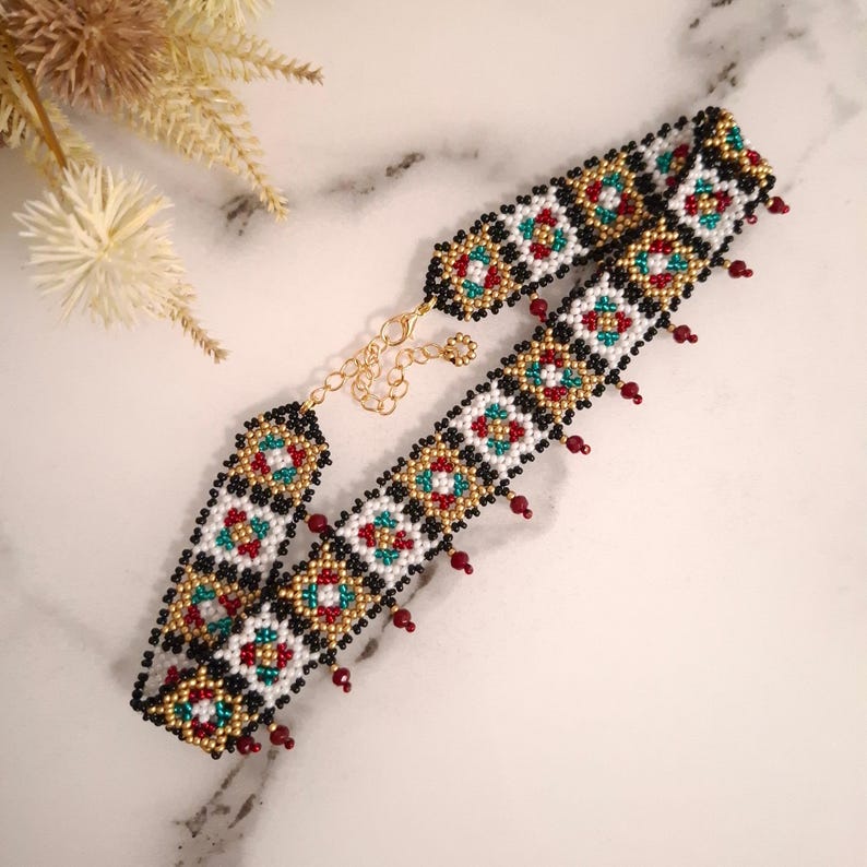 K&ouml;nnte beinhalten: Eine Halskette mit Perlen im Choker-Stil mit einem geometrischen Muster in Schwarz, Wei&szlig;, Gold, Rot und T&uuml;rkis. Die Halskette hat einen goldfarbenen Kettenverschluss und kleine rote Perlen, die unten h&auml;ngen.