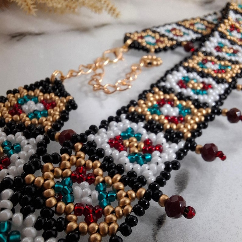 K&ouml;nnte beinhalten: Eine Halskette mit Perlenbesatz, mit einer goldfarbenen Kette und einem Verschluss. Die Halskette hat ein geometrisches Muster aus schwarzen, wei&szlig;en, goldenen, roten und t&uuml;rkisfarbenen Perlen. Kleine, dunkelrote Perlen h&auml;ngen von der Unterseite der Halskette.